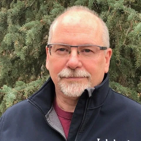 Middle-aged man wearing glasses and a navy jacket, smiling in front of evergreen trees.