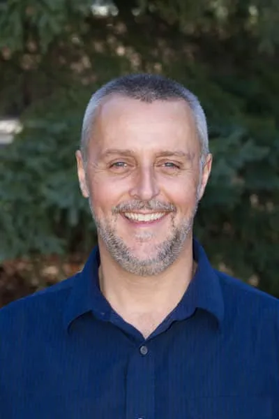 Smiling middle-aged man with short gray hair and a beard, wearing a blue collared shirt, standing outdoors with green foliage in the background.