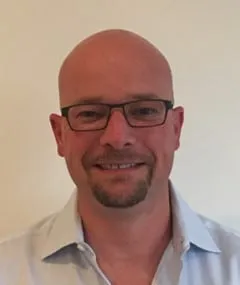 Smiling man with a bald head and glasses, wearing a light blue collared shirt, against a neutral background.