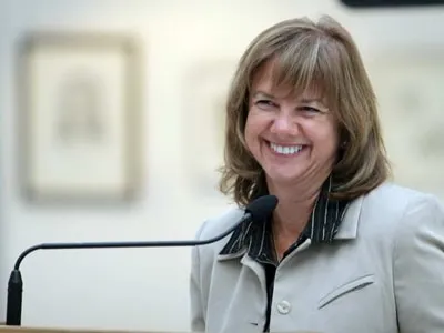 Smiling woman in a professional setting, speaking at a podium with a microphone, showcasing a confident and engaging demeanor. Background features blurred artwork, suggesting an art gallery or exhibition.