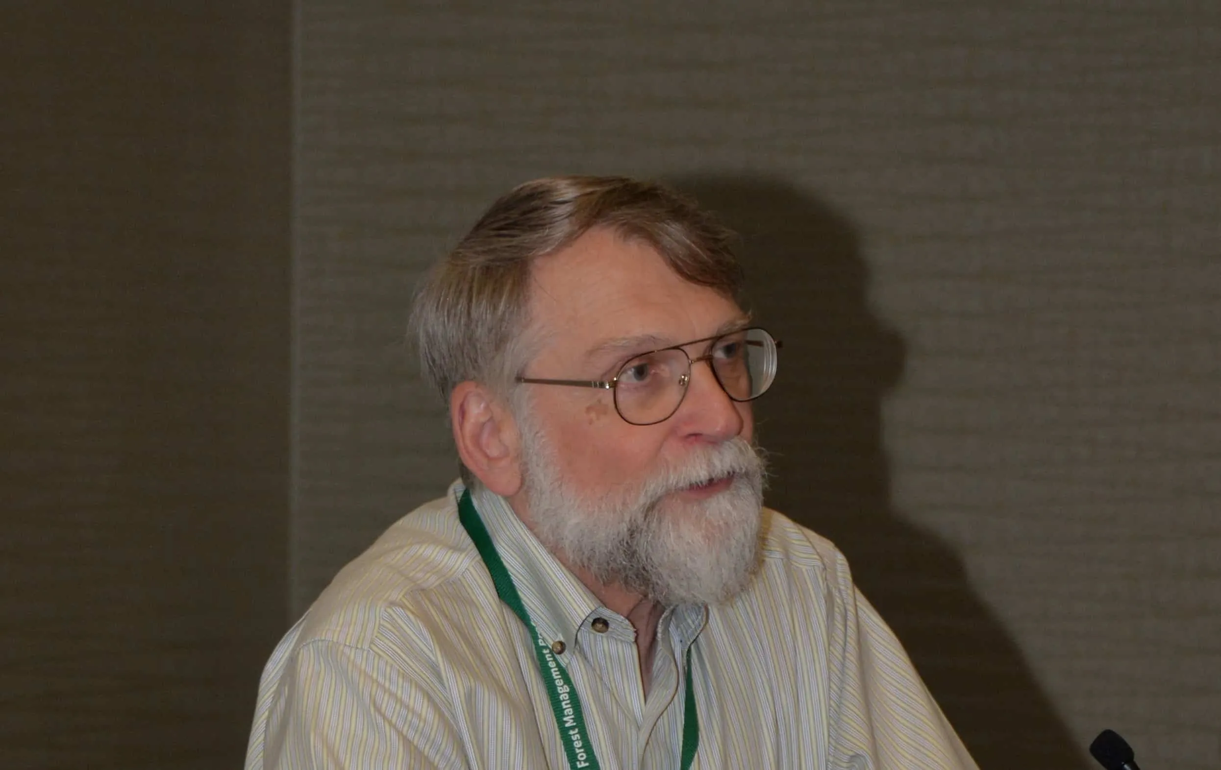 Older man with glasses and a beard engaged in a discussion, wearing a striped shirt and a green lanyard at a conference setting.