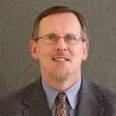 Professional headshot of a middle-aged man with glasses, wearing a suit and tie, against a gray background.