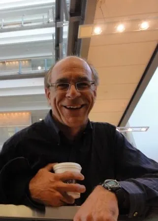 Smiling man holding a coffee cup, wearing glasses and a dark shirt, standing indoors with modern architecture in the background.