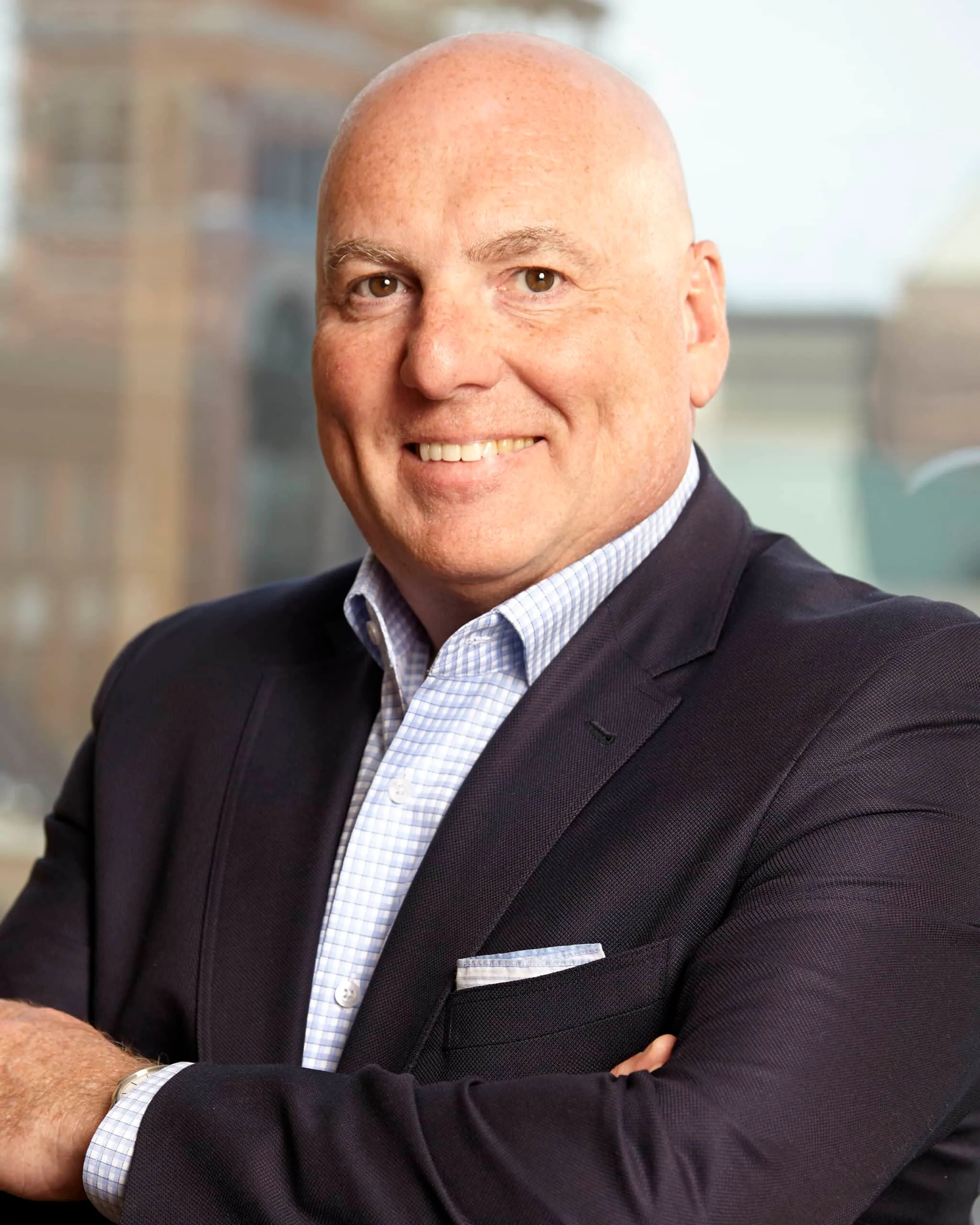 Professional headshot of a smiling bald man in a dark blazer over a light checkered shirt, posing with arms crossed in front of a blurred urban background. Ideal for business profiles and corporate websites.