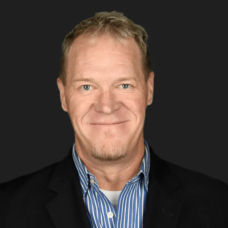 Professional headshot of a smiling man with short blonde hair and a goatee, wearing a black blazer over a blue and white striped shirt, set against a dark background.