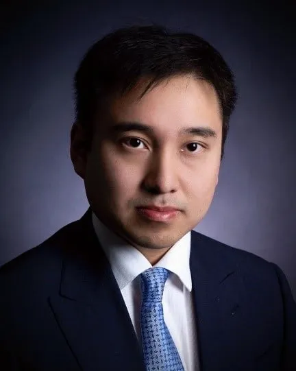 Portrait of a professional man in a dark suit and light blue tie, with a neutral expression against a softly lit background. Ideal for business or corporate contexts.