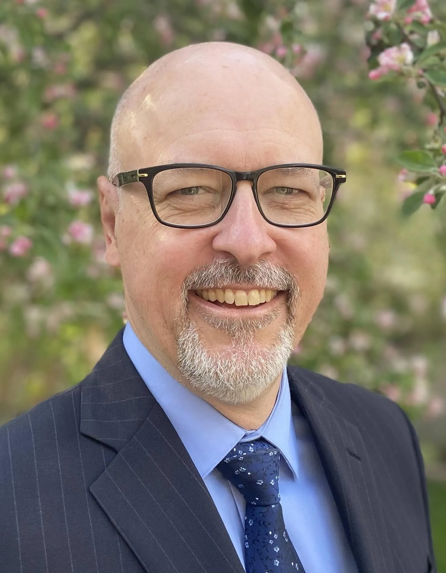 Smiling middle-aged man in a suit and tie, standing outdoors with a blurred background of pink flowering trees, showcasing a professional and approachable demeanor.
