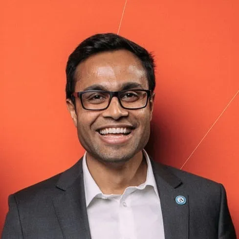 Smiling professional man in a suit and glasses against an orange background, showcasing a friendly and approachable demeanor.