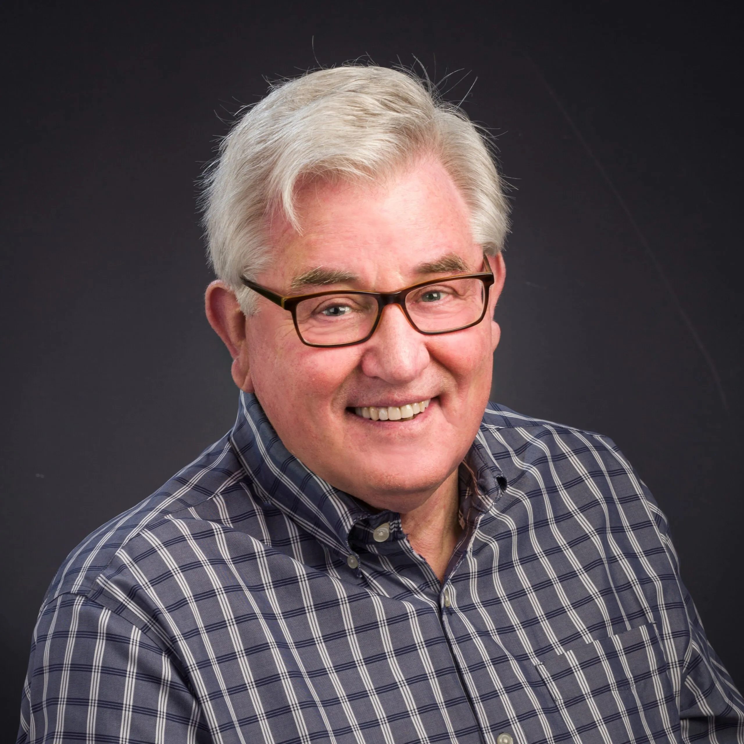 Smiling older man with gray hair and glasses wearing a plaid shirt, posing against a dark background.