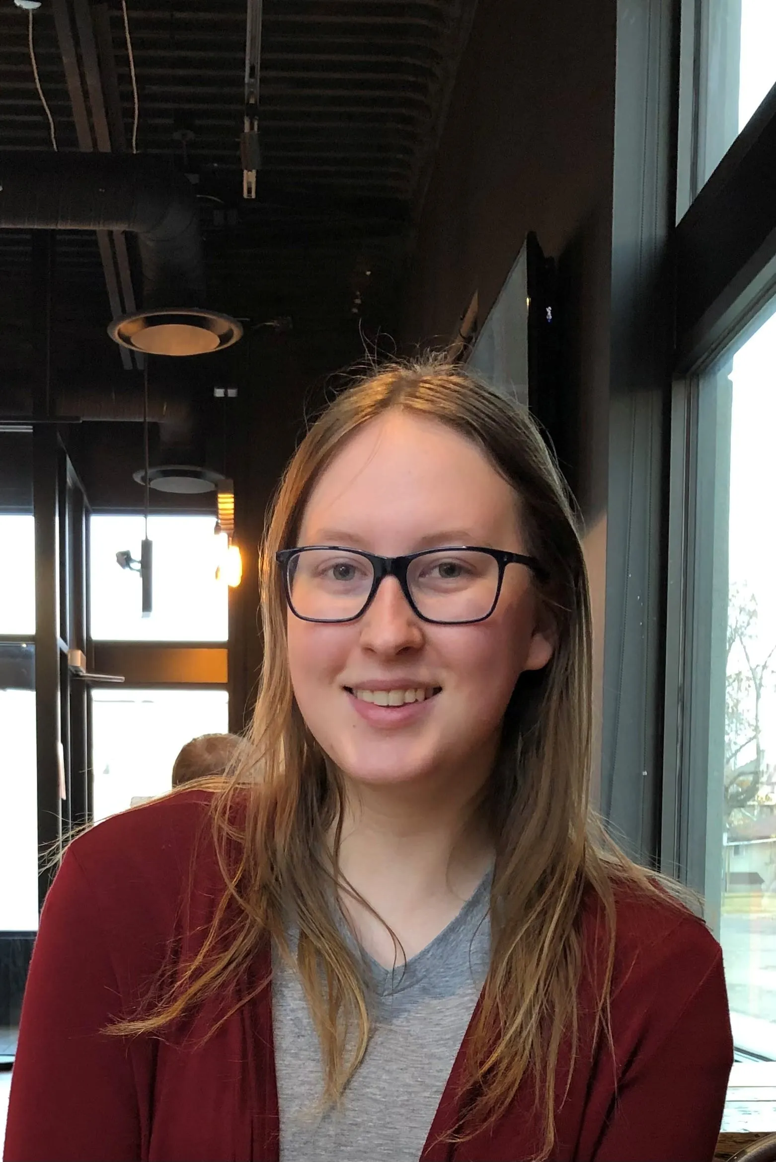 Young woman with long hair wearing glasses and a maroon cardigan, smiling in a cafe setting with large windows and modern interior design.