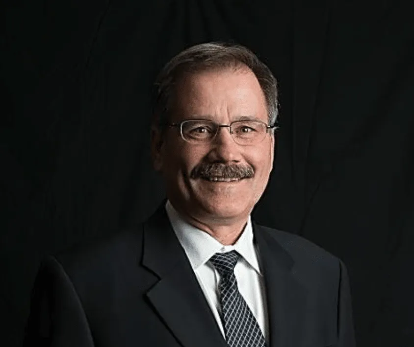 Professional portrait of a middle-aged man with glasses and a mustache, wearing a black suit and tie, set against a dark background. Ideal for business profiles or corporate websites.