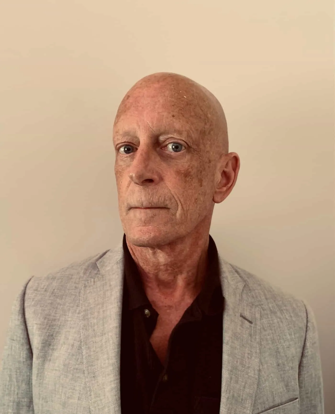 Portrait of a mature man with a bald head and freckles, wearing a light gray blazer over a black shirt, against a neutral background.