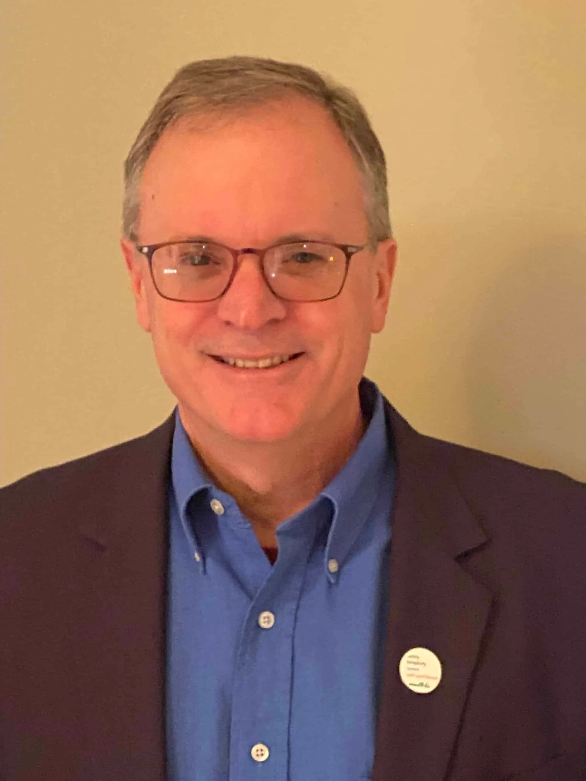 Smiling middle-aged man wearing glasses and a blue shirt under a dark blazer, standing against a neutral background.