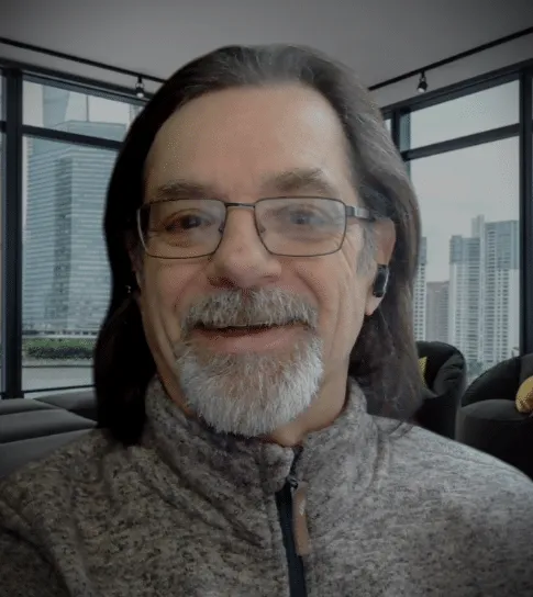 Smiling middle-aged man with glasses and a gray beard, wearing a zip-up sweater, sitting in a modern indoor setting with large windows showcasing a cityscape in the background.