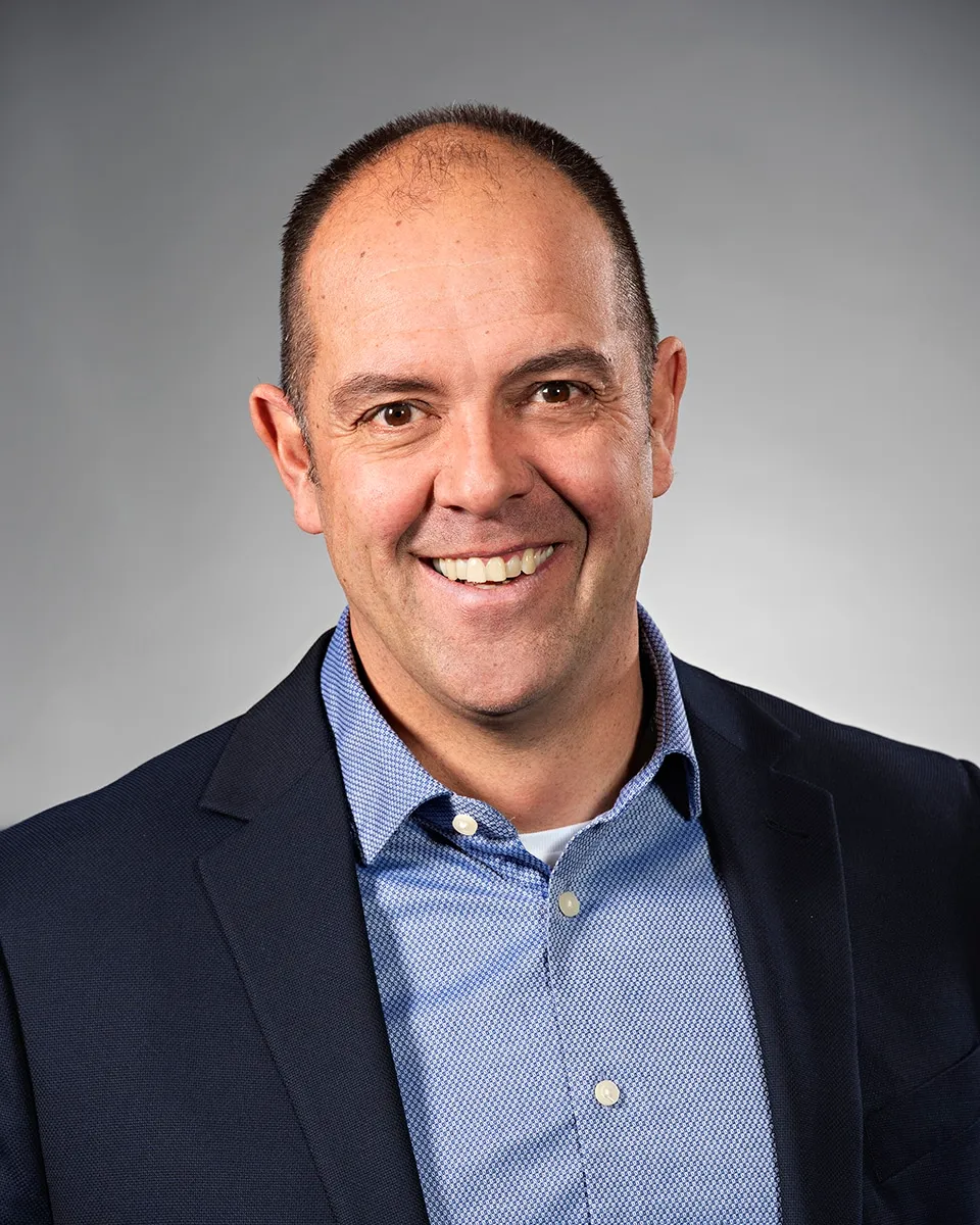 Professional headshot of a smiling man wearing a dark blazer over a patterned blue shirt, set against a neutral gray background. Ideal for business profiles or corporate communications.