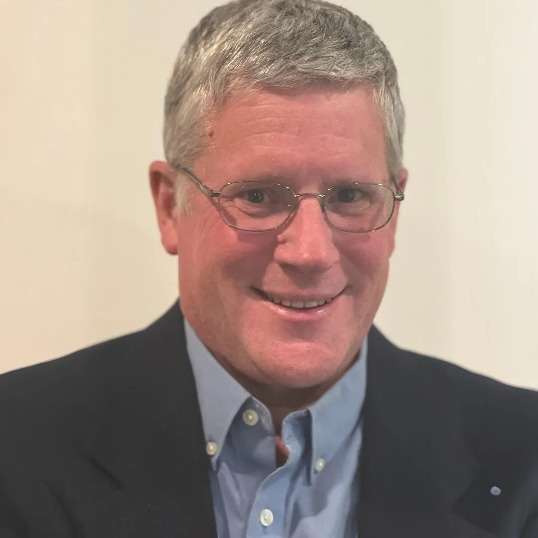 Professional headshot of a smiling man with short gray hair, wearing glasses, a blue collared shirt, and a dark blazer, set against a neutral background.