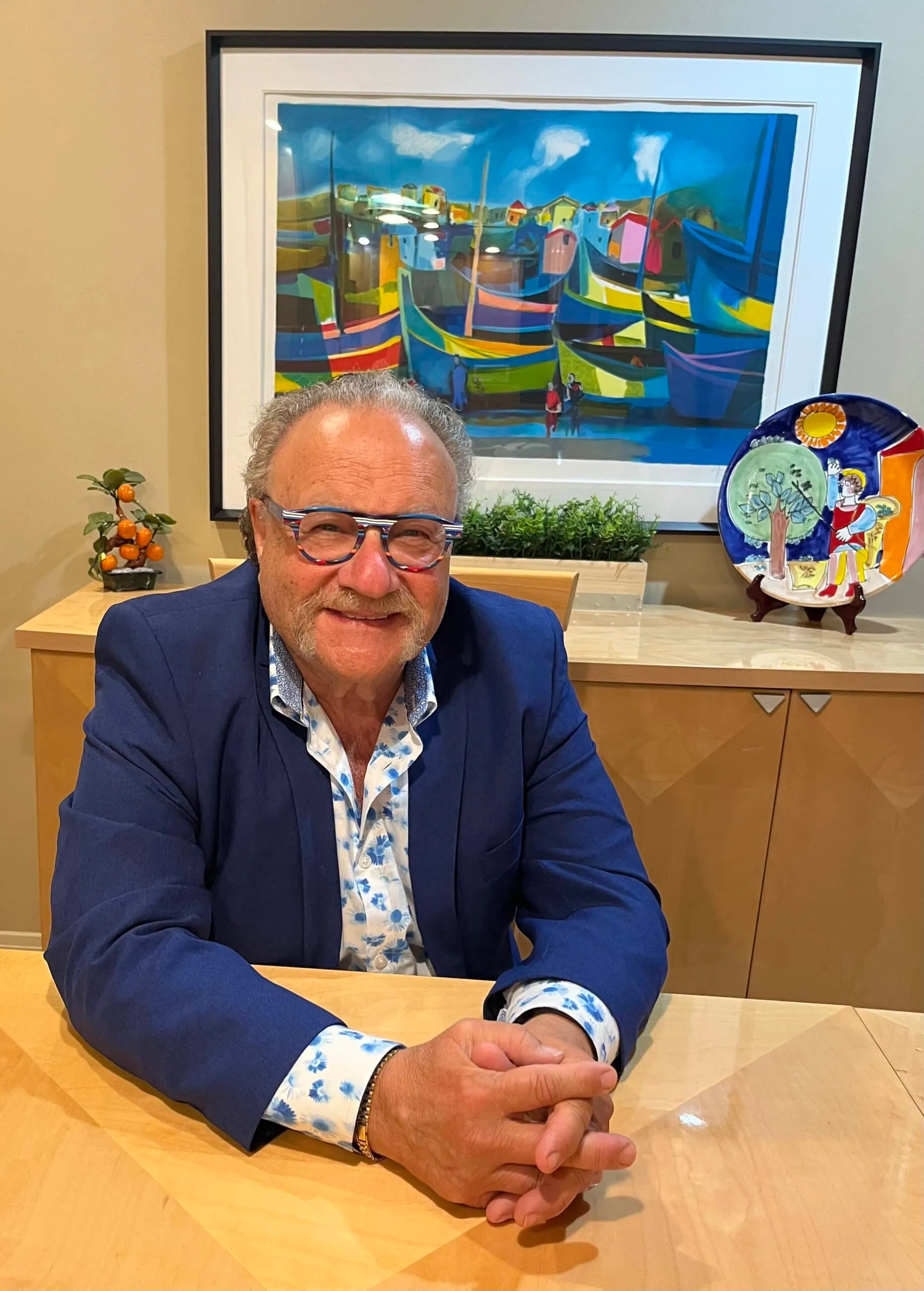 Smiling man with glasses seated at a wooden table, wearing a blue blazer over a floral shirt, with a colorful painting of boats in the background and decorative items on the table.