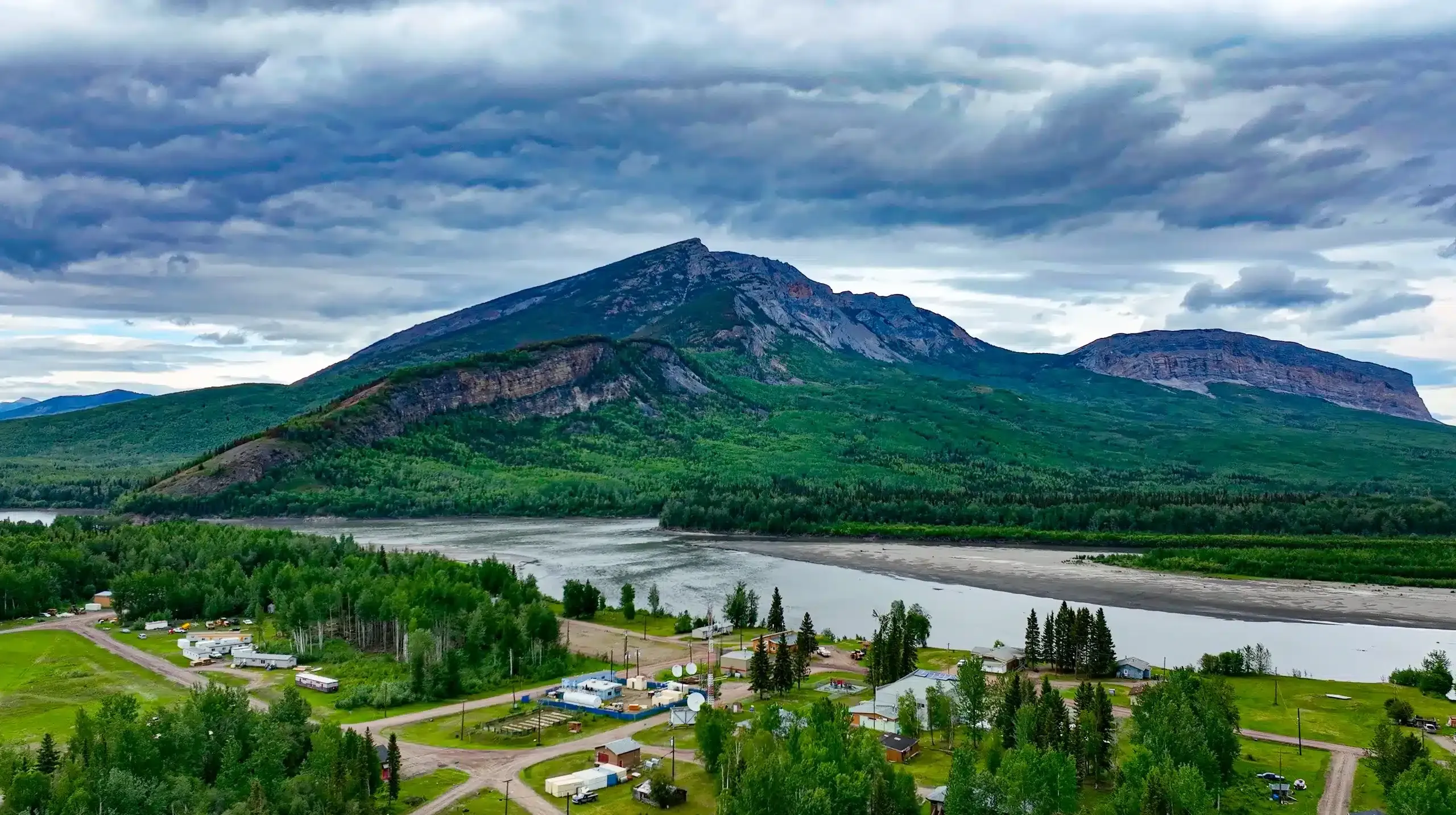 Aerial view of a scenic landscape featuring a large mountain surrounded by lush greenery, with a river flowing nearby and small residential structures visible in the foreground. The sky is overcast, adding a dramatic effect to the natural beauty of the area.
