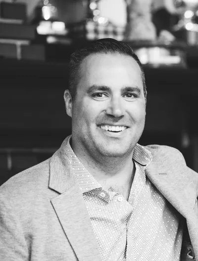 Smiling man in a light gray blazer and patterned shirt, standing in a cozy indoor setting with blurred trophies and decorations in the background.