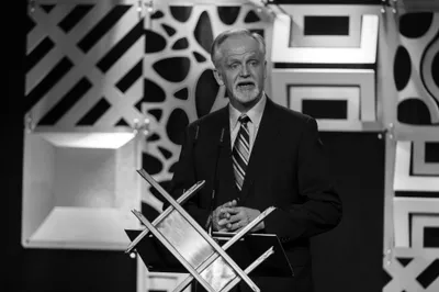 A man in a suit delivers a speech at a podium, with a modern, abstract backdrop featuring geometric patterns. The image is in black and white, emphasizing the formal setting of the event.