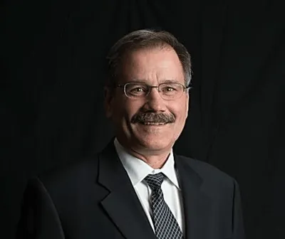 Professional portrait of a middle-aged man with glasses and a mustache, wearing a black suit and tie, set against a dark background. Ideal for business profiles or corporate websites.