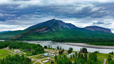 Aerial view of a scenic landscape featuring a large mountain surrounded by lush greenery, with a river flowing nearby and small residential structures visible in the foreground. The sky is overcast, adding a dramatic effect to the natural beauty of the area.